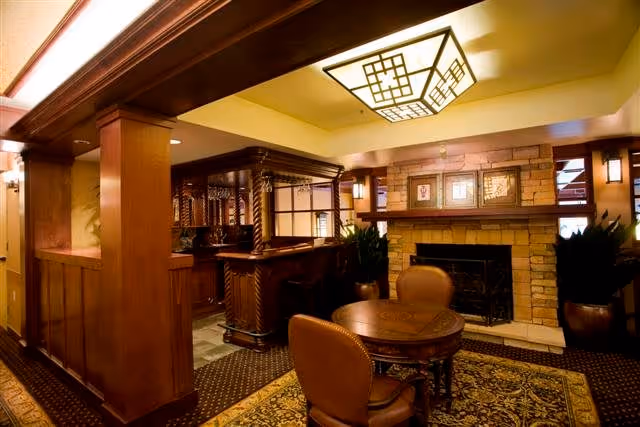 A cozy interior lounge area featuring a wooden bar with ornate detailing, a round wooden table with four leather chairs, a stone fireplace with framed artwork above it, and warm lighting from a decorative ceiling fixture and wall sconces.