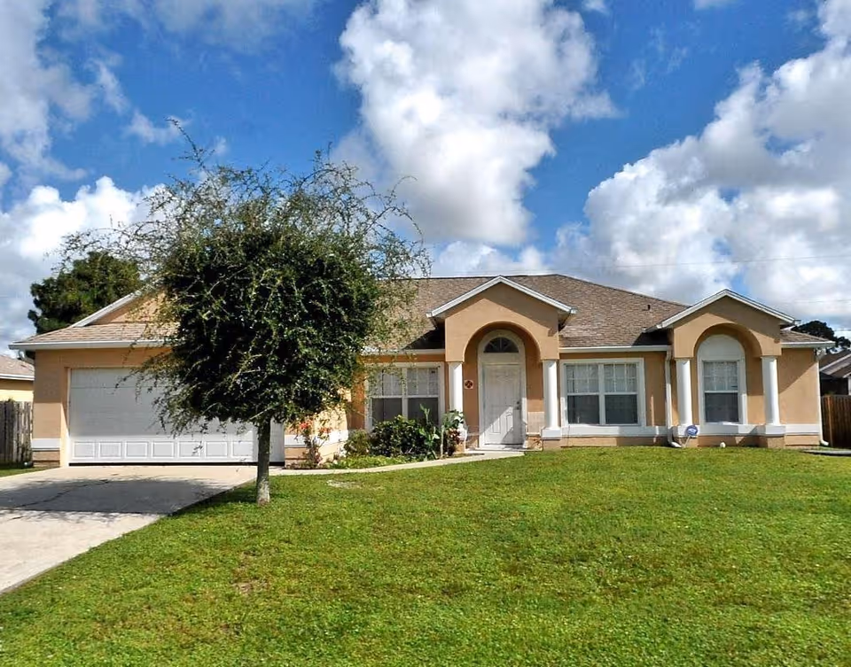 Single-story peach-colored house with an attached garage, front lawn and a small tree under a partly cloudy sky.
