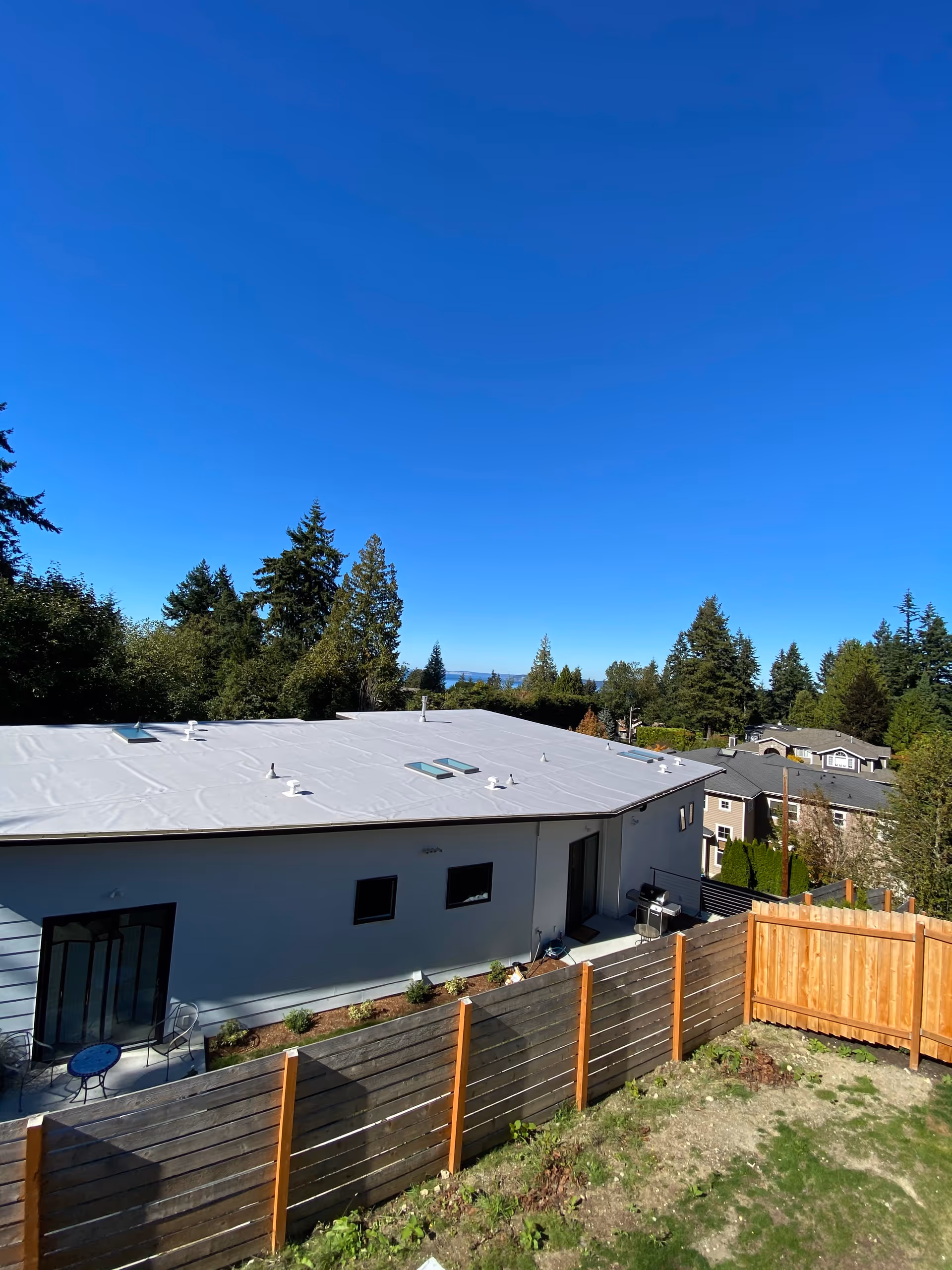 View of a single-story modern house with a white flat roof, wooden fence and backyard under a clear blue sky.