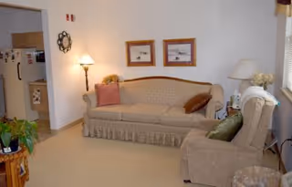 A cozy living room area featuring a beige sofa with two decorative pillows, a matching armchair with a green pillow, a side table with a lamp and flowers, two framed pictures on the wall, and a glimpse into an adjacent kitchen area with a refrigerator and cabinets.