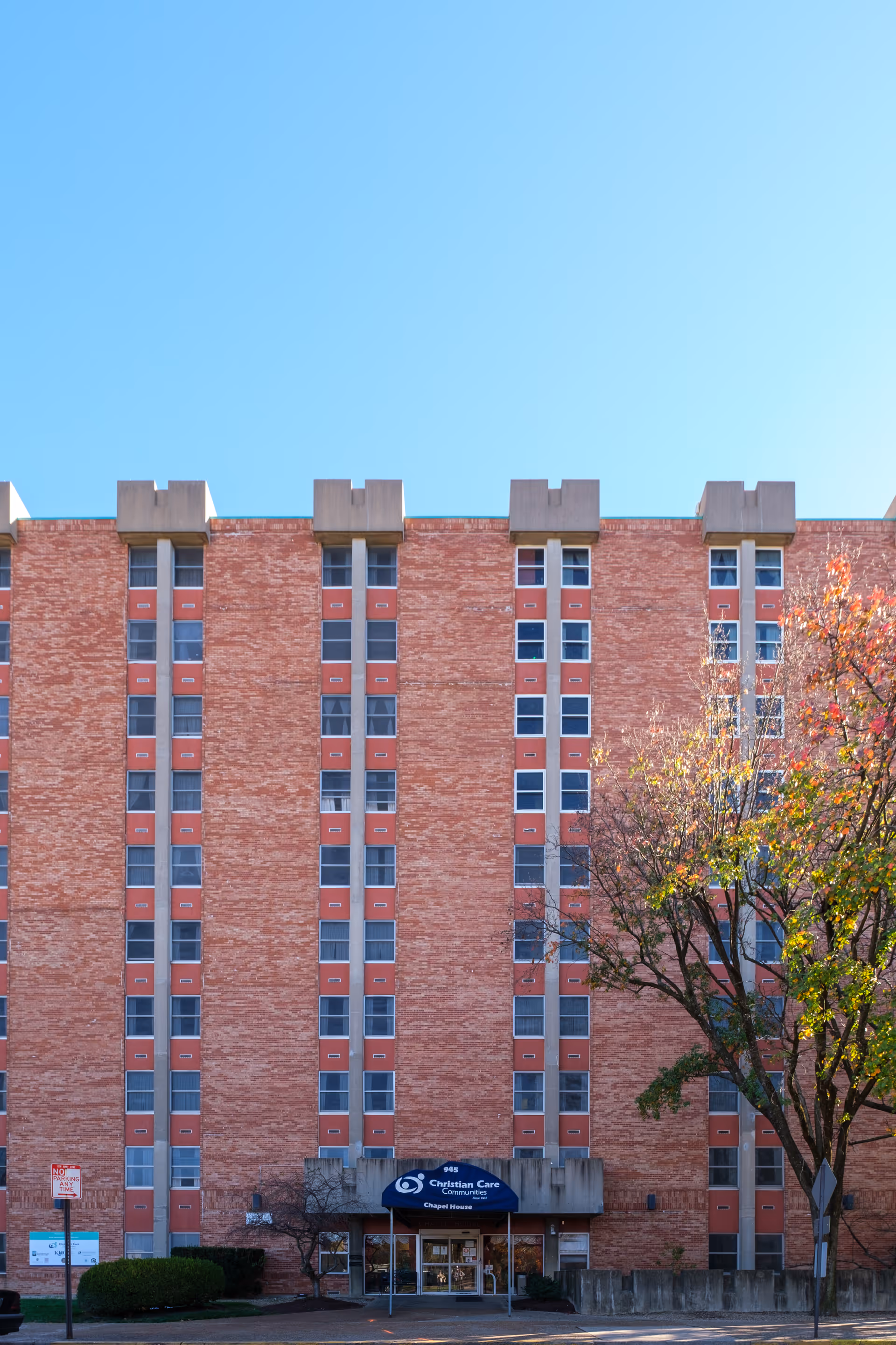 Exterior front view of a multi-story brick building with vertical rows of windows and a blue awning above the entrance that reads 'Christian Care Communities Chapel House'. A tree with autumn foliage is visible on the right side under a clear blue sky.