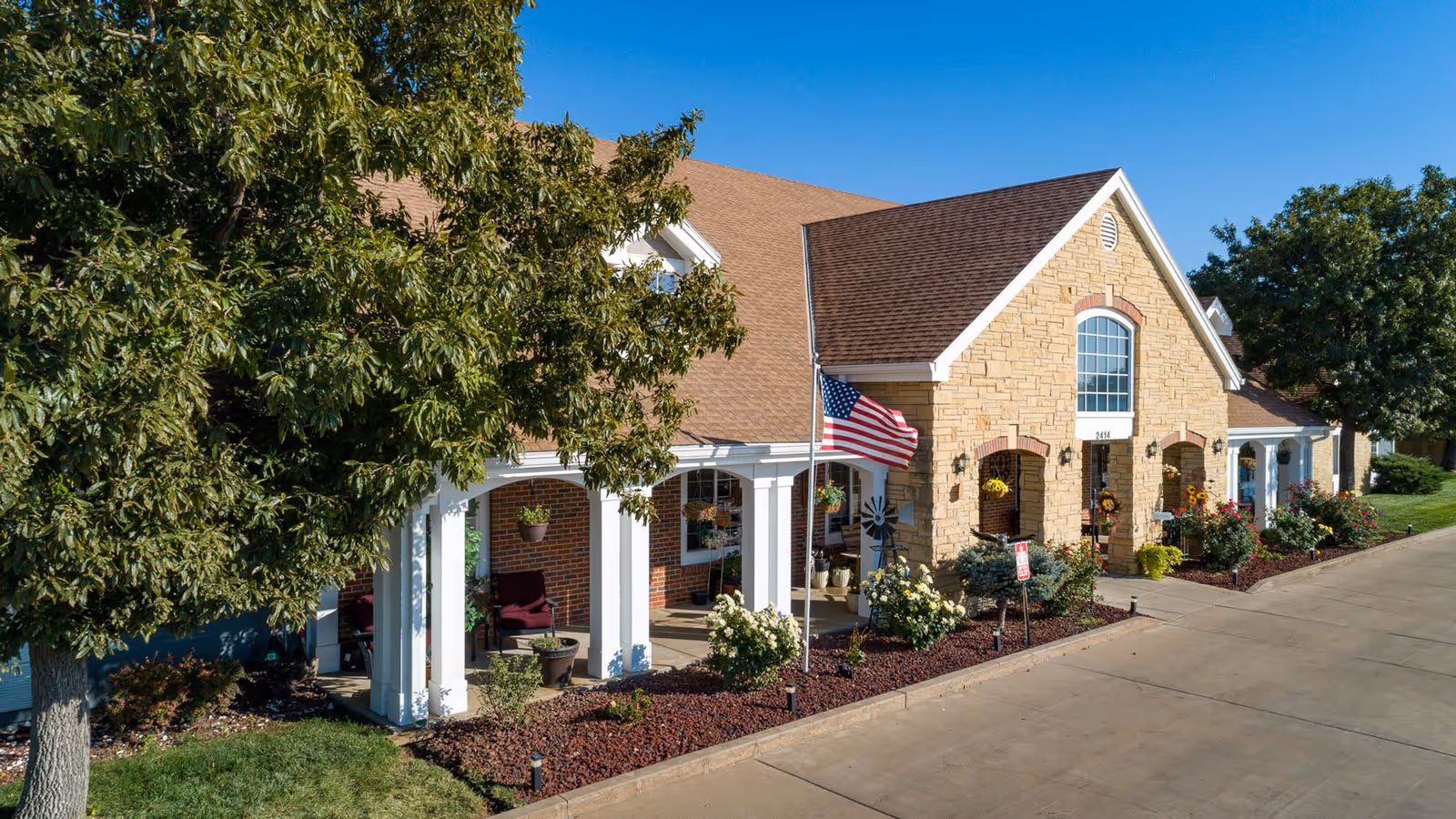 Exterior view of Homestead Assisted Living of Garden City showing a single-story building with a stone and brick facade, a brown shingled roof, white columns supporting a covered porch, hanging flower pots, an American flag, and landscaped flower beds along the sidewalk under a clear blue sky.