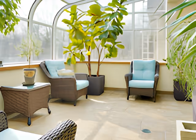 Bright indoor seating area with wicker chairs featuring light blue cushions, a small wicker side table, and several large potted plants near large windows with curved frames letting in natural light.