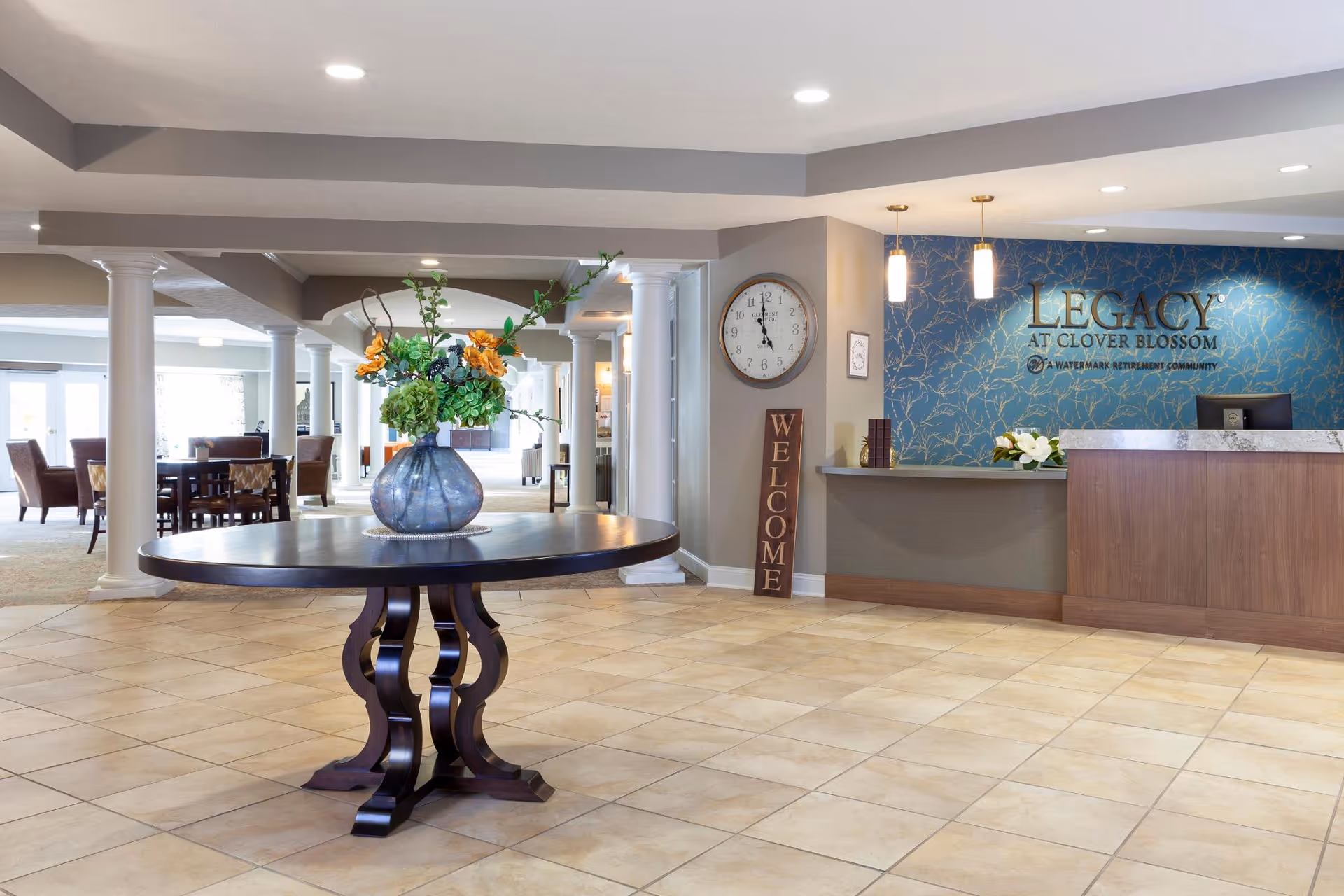 Spacious lobby area of Legacy at Clover Blossom featuring a round wooden table with a large vase of flowers in the center. To the right is a reception desk with a blue patterned wall behind it displaying the facility's name. A clock and a vertical 'WELCOME' sign are on the wall near the desk. The background shows seating areas with chairs and tables, supported by white columns.