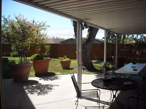 Shaded outdoor patio with metal tables and chairs, potted plants, and a grassy yard.
