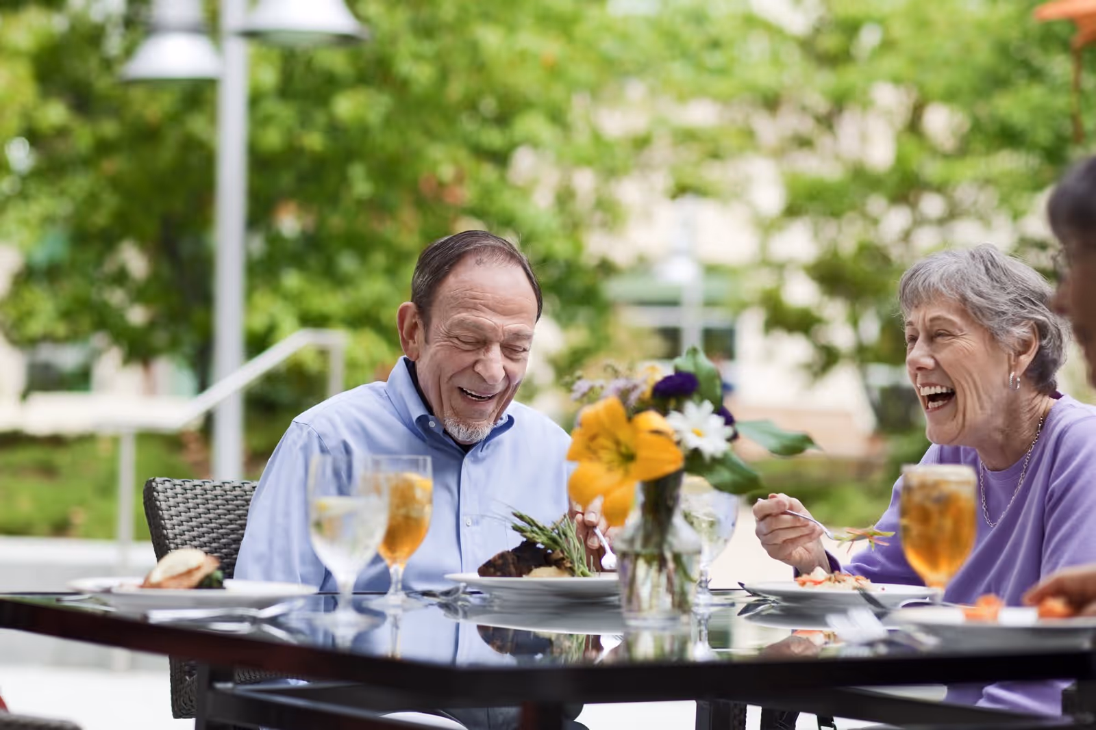 Two older adults laughing and eating at an outdoor dining table with drinks and a flower centerpiece.