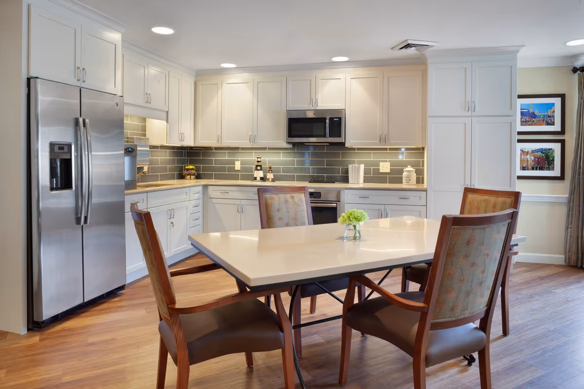 A modern kitchen with white cabinetry, stainless steel refrigerator, microwave, and oven. There is a rectangular dining table with four wooden chairs around it, and a small vase with green flowers on the table. The kitchen has a gray tile backsplash and wooden flooring.
