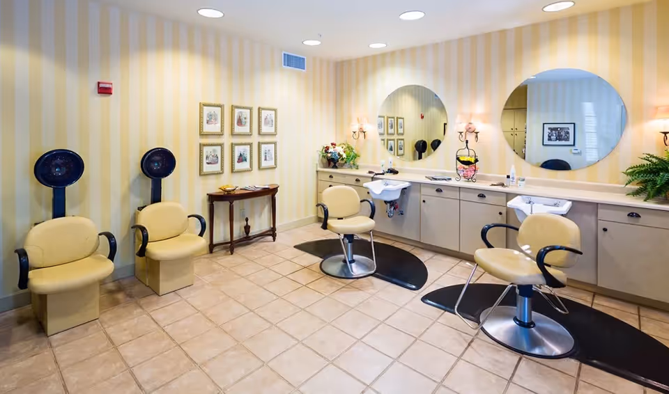 Interior of a hair salon area in an independent living community featuring two beige salon chairs with black armrests in front of sinks and large round mirrors mounted on a yellow striped wall. Two beige chairs with black hair dryers are positioned against the left wall. The floor is tiled, and there is a small wooden table with framed pictures above it on the wall.