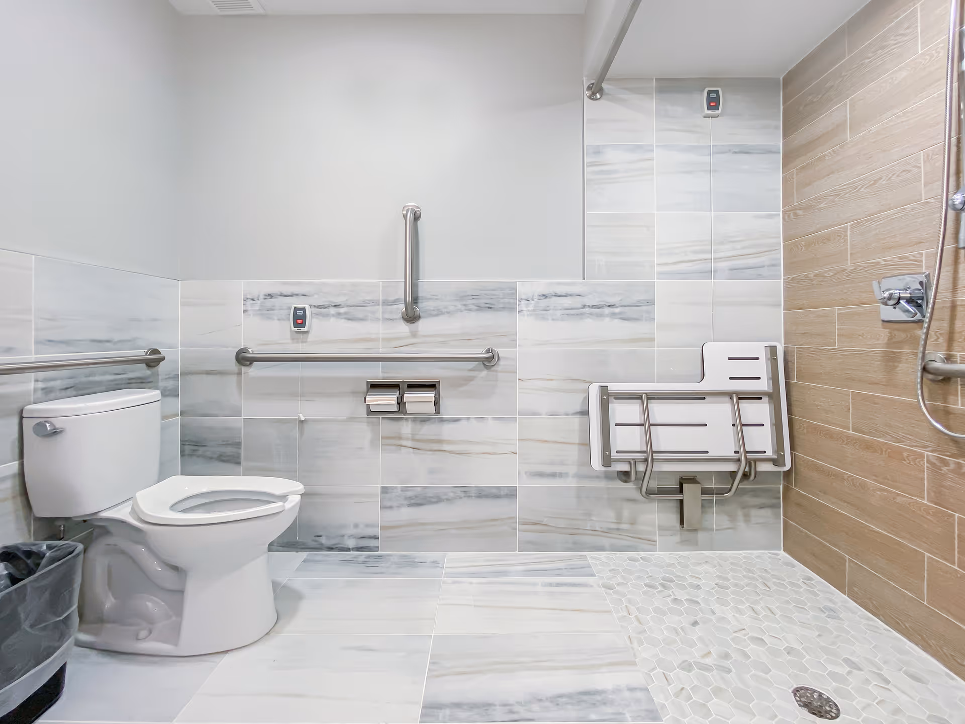 A modern accessible bathroom featuring a white toilet with grab bars on the walls, a foldable shower seat, and a walk-in shower area with a handheld showerhead. The walls are tiled with a combination of light marble and wood-like tiles, and the floor has matching marble and pebble-style tiles.
