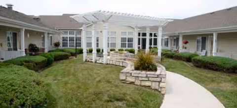Outdoor courtyard with a white pergola, stone walls, and trimmed shrubs around beige single-story buildings.