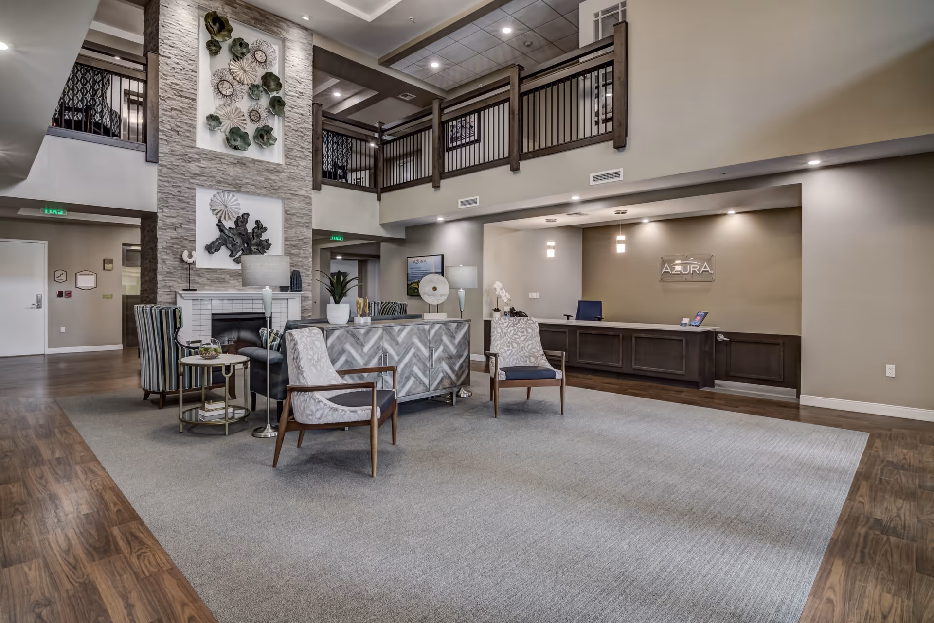 Spacious and modern assisted living facility lobby with a high ceiling and upper balcony railing. The area features a seating arrangement with patterned chairs and a small table on a large carpet. A stone fireplace with decorative wall art is centered on one wall. The reception desk is visible on the right side with a sign that reads 'AZURA'. The flooring is a combination of wood and carpet, and the lighting is soft and warm.