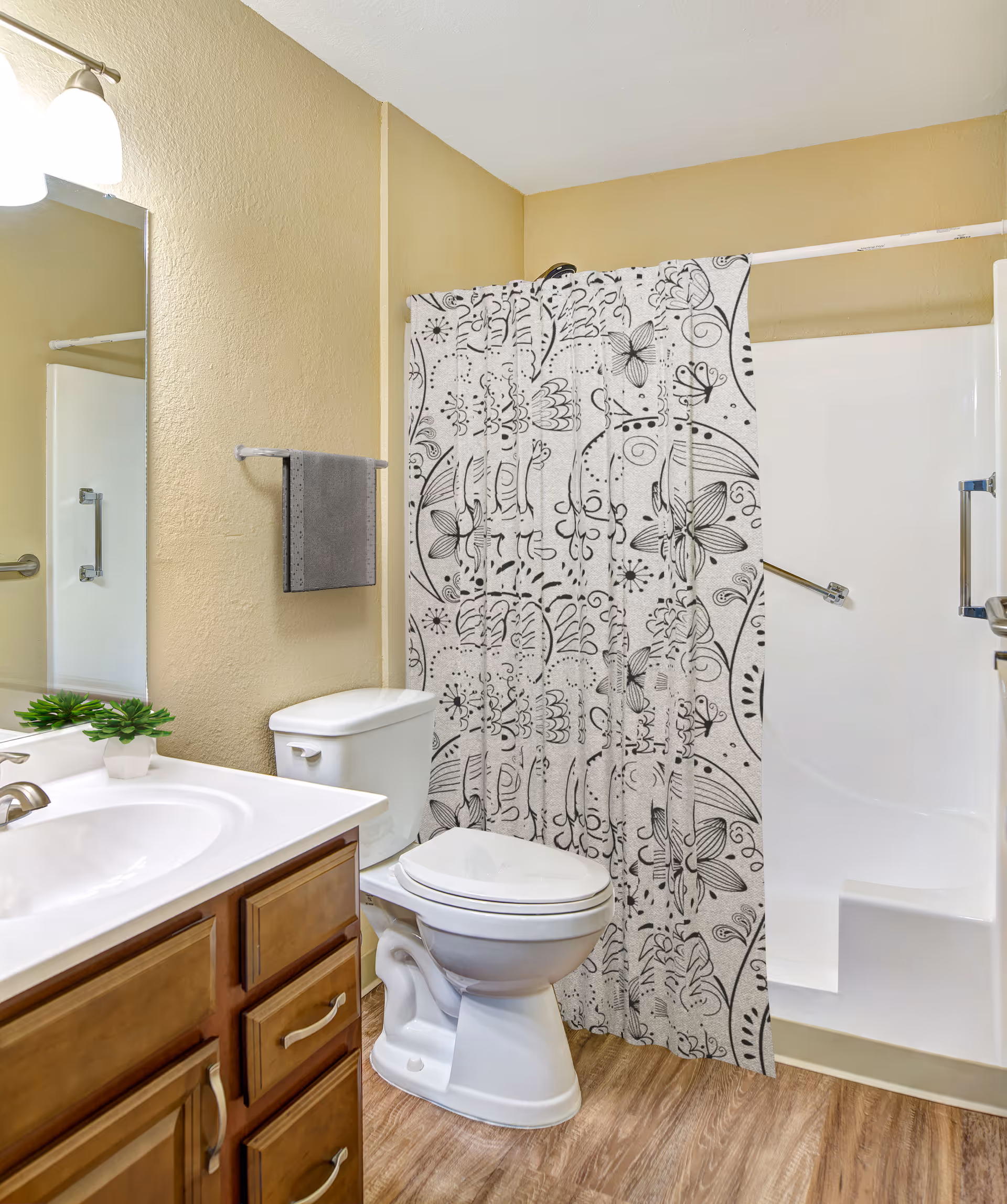 A clean bathroom with a white toilet, a wooden vanity with a white sink, a small green plant on the counter, a large mirror, a gray towel hanging on a towel rack, and a shower with a white and black patterned curtain.