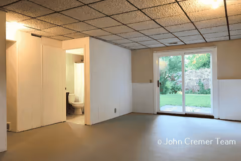 Basement living area with a tiled drop ceiling, sliding glass door to a backyard, and an open doorway revealing a bathroom.