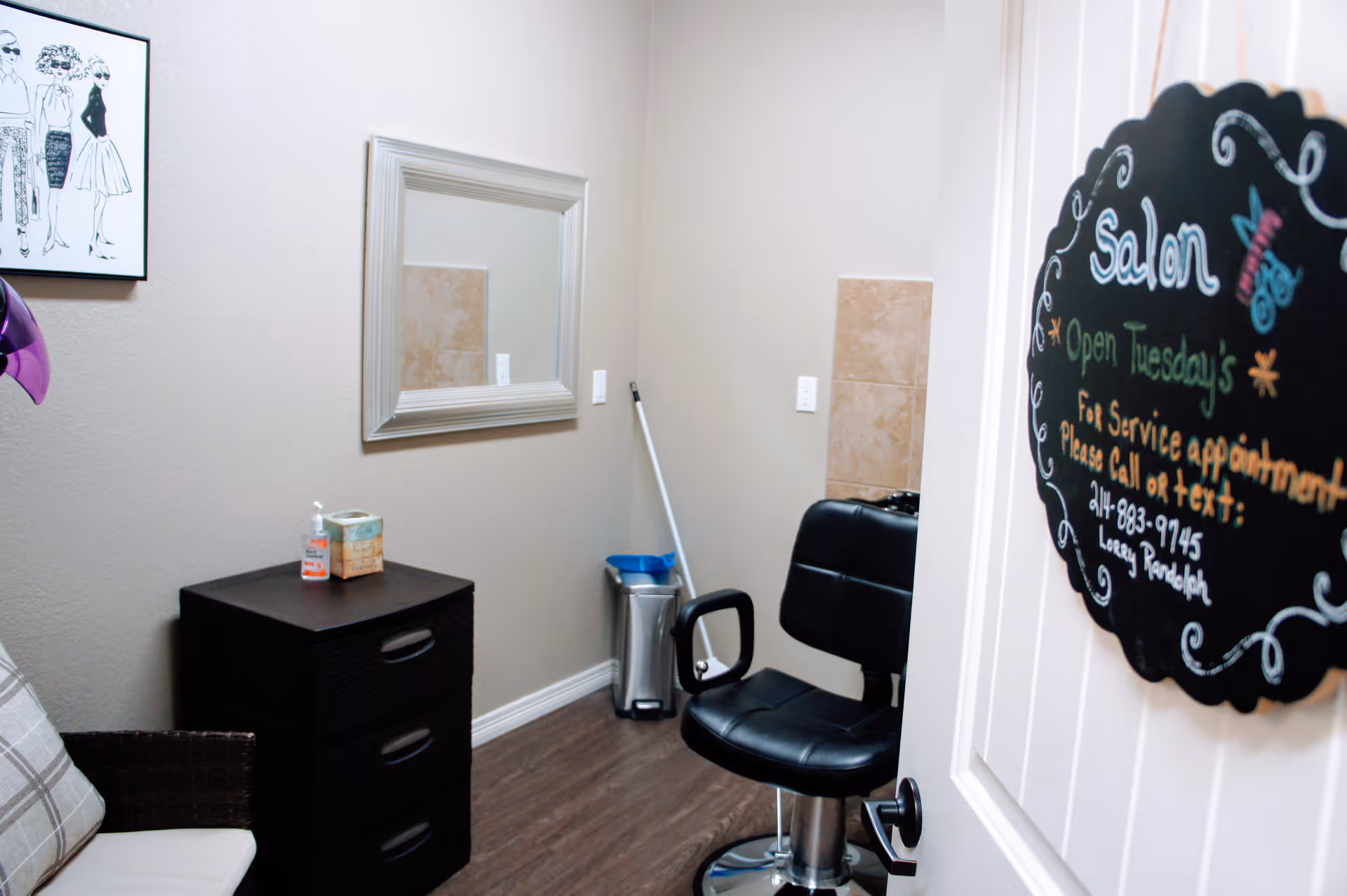 Interior view of a small salon room with a black salon chair, a black three-drawer cabinet with hand sanitizer and a tissue box on top, a wall mirror, a mop and trash can in the corner, and a decorative sign on the door indicating salon service appointment details.