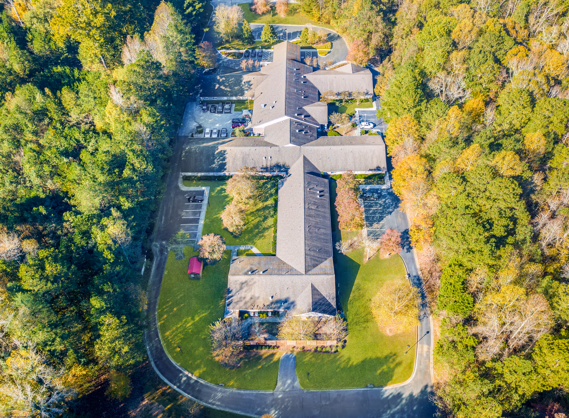Aerial view of a cross-shaped residential facility building with surrounding lawns, parking areas, and dense trees.