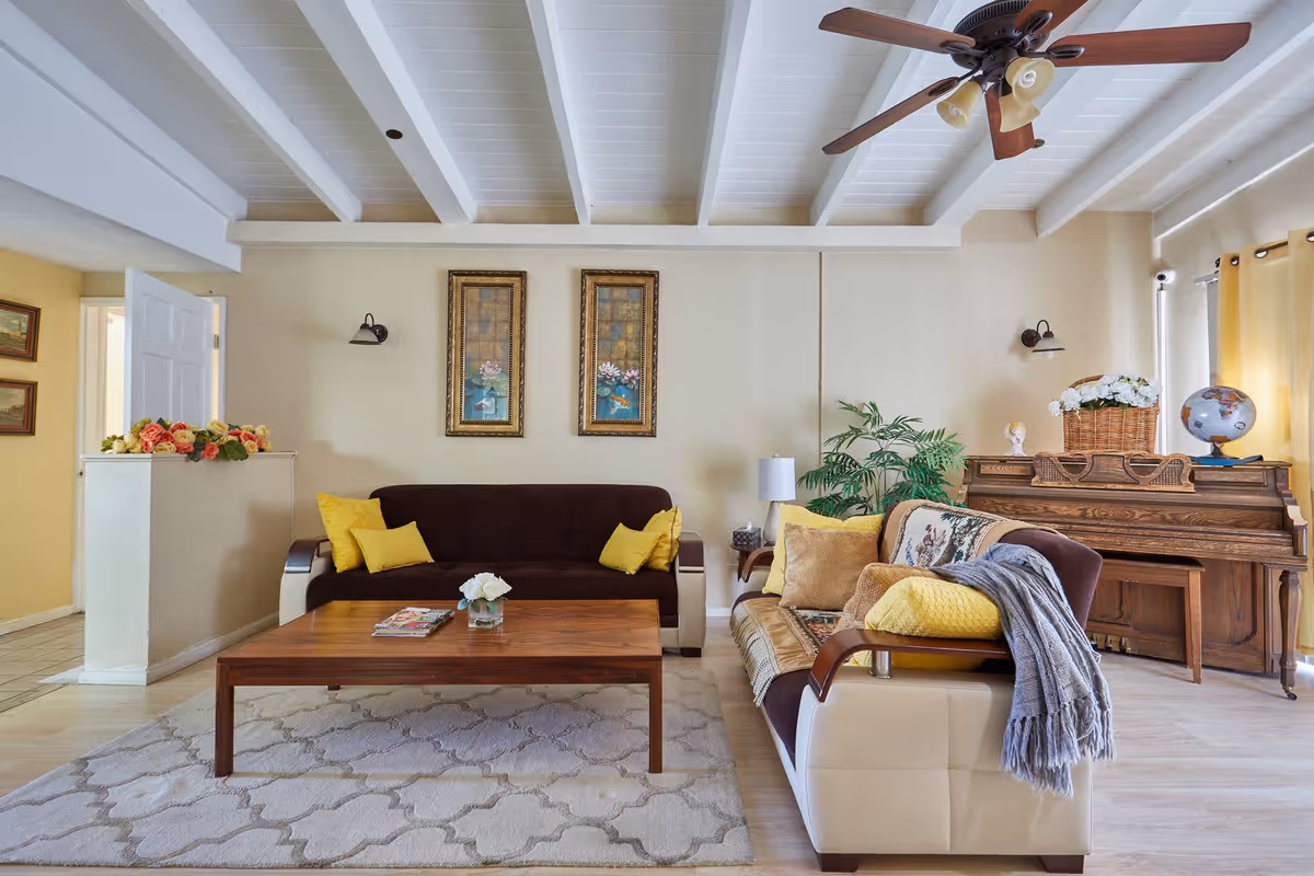 A cozy living room with two sofas adorned with yellow and beige cushions, a wooden coffee table with a small flower vase and magazines, a wooden piano with a basket of flowers and a globe on top, framed artwork on the wall, a ceiling fan with lights, and a potted plant near the corner.