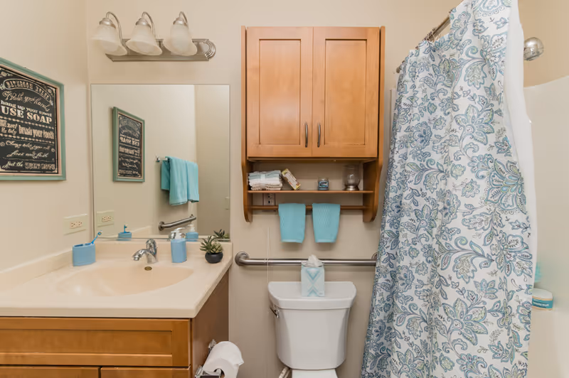 A bathroom with a beige countertop and sink, a large mirror above the sink, and a wooden cabinet mounted on the wall above a white toilet. There are two turquoise hand towels hanging on a rack under the cabinet, a patterned shower curtain with blue and green designs, and a small plant on the countertop. A framed sign with bathroom rules is visible on the wall next to the mirror.
