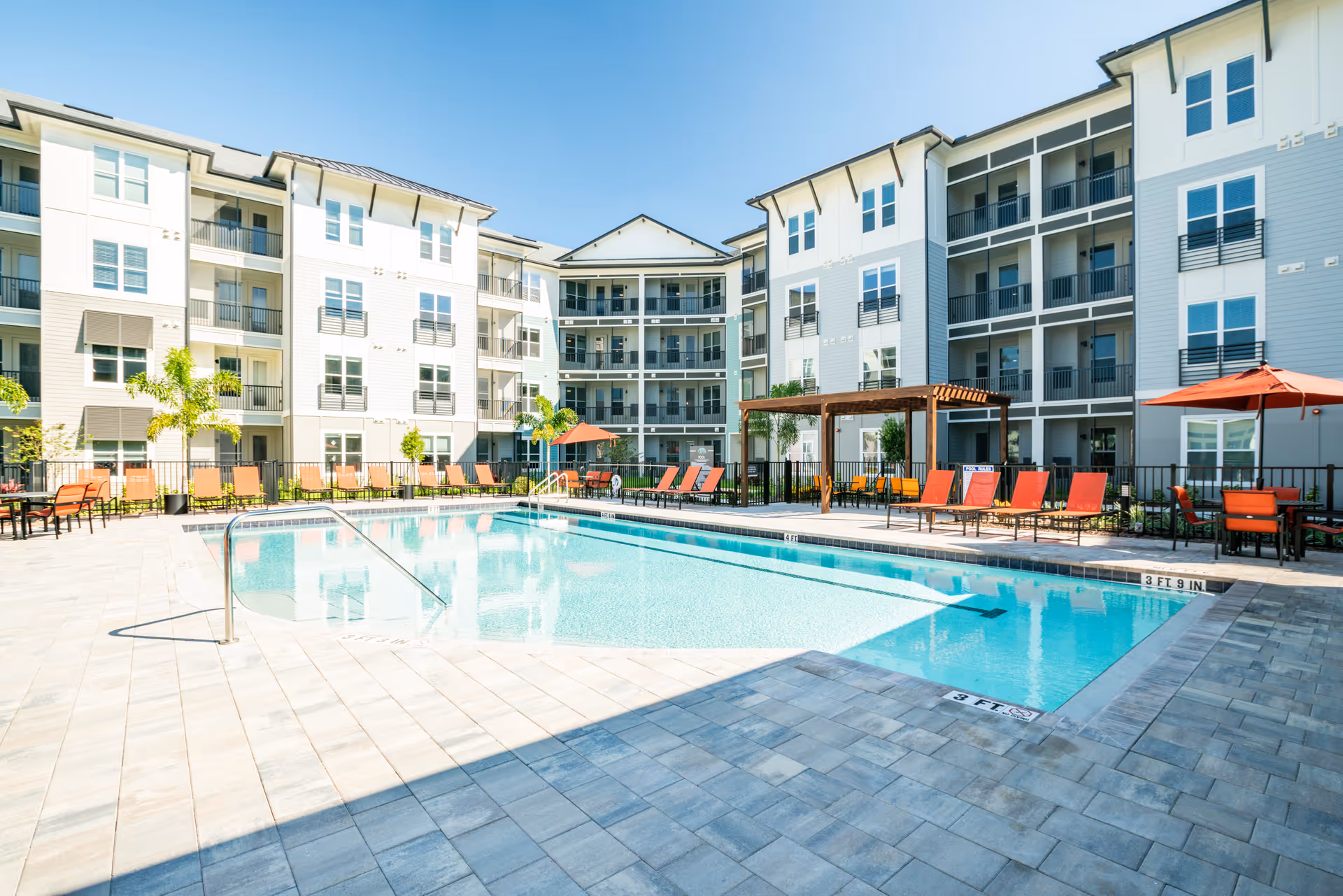 Outdoor courtyard with a rectangular swimming pool surrounded by lounge chairs, umbrellas, and multi-story apartment buildings.