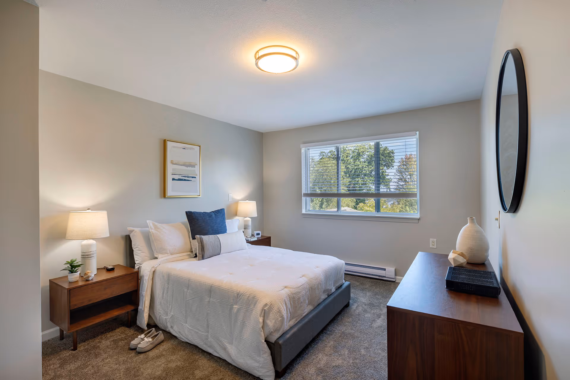 A neatly made bed with white bedding and blue accent pillows in a bedroom. There are two wooden nightstands with lamps on either side of the bed. A pair of slippers is placed on the carpeted floor next to one nightstand. A large window with blinds lets in natural light, showing green trees outside. A wooden dresser with decorative items and a round mirror above it is on the right wall.