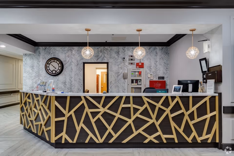 Reception desk area with a modern design featuring a dark wood front with light wood geometric patterns. Three spherical pendant lights hang from the ceiling above the desk. Behind the desk is a wall with a textured gray and white pattern, a large round clock, and an open doorway leading to another room. Various office equipment and informational materials are on the desk.