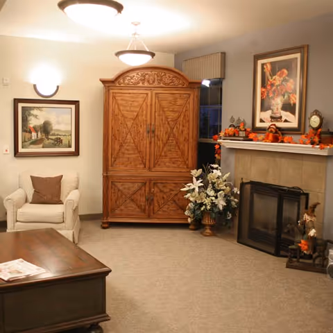 A cozy living room with a beige armchair with a brown cushion, a large wooden armoire with intricate carvings, a fireplace decorated with autumn-themed garlands and a framed floral painting above it, a vase with white flowers, and a wooden coffee table with a newspaper on it.