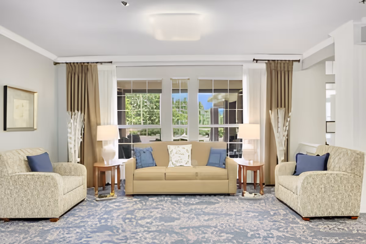 A bright living room with a beige sofa in the center flanked by two matching armchairs, each with a blue cushion. There are two wooden side tables with lamps on either side of the sofa. Large windows with brown and white curtains allow natural light to fill the room, and a blue patterned carpet covers the floor.