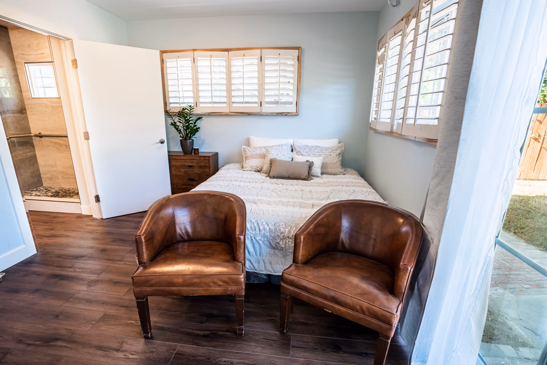 A bright bedroom with a bed covered in white bedding and multiple pillows against a wall with windows featuring white shutters. Two brown leather chairs are positioned in front of the bed on a wooden floor. To the left, an open door reveals a bathroom with a glass shower. On the right, a large window with white curtains looks out onto a grassy outdoor area.