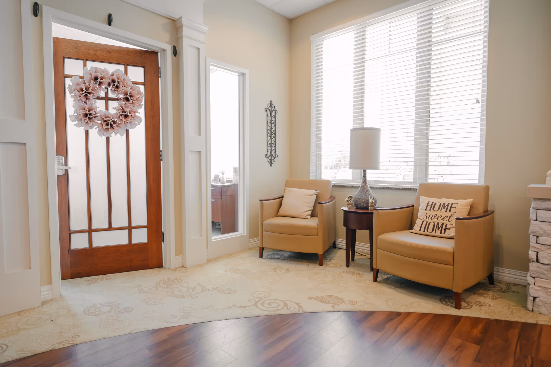 A cozy sitting area with two tan armchairs, one with a pillow that says 'Home sweet Home', a small round wooden table with a lamp and decorative items, next to a large window with white blinds. To the left is a wooden door with glass panels and a pink floral wreath, and a narrow window beside it. The floor has a combination of light patterned carpet and dark wood.