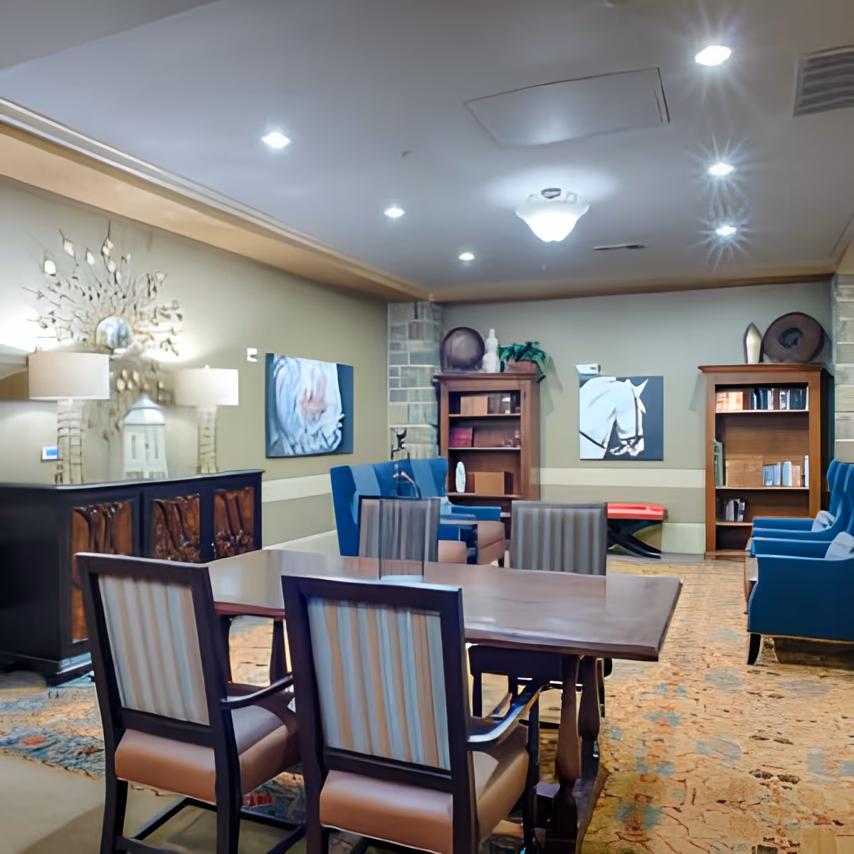 A well-lit common area in a senior living facility featuring a wooden table with four striped upholstered chairs in the foreground. The room has blue armchairs, wooden bookshelves, decorative artwork on the walls, and a sideboard with two lamps and a decorative mirror above it. The carpet has a patterned design, and the ceiling has recessed lighting.