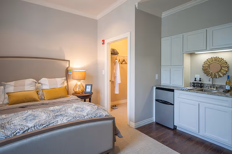 A cozy bedroom in an assisted living facility featuring a neatly made bed with patterned bedding and yellow accent pillows. A bedside table with a lamp and a framed picture is next to the bed. There is an open door revealing a walk-in closet with hanging clothes and shelves. To the right, a small kitchenette area with white cabinets, a sink, a mini refrigerator, and decorative items including a sunburst mirror and a bottle of wine is visible.