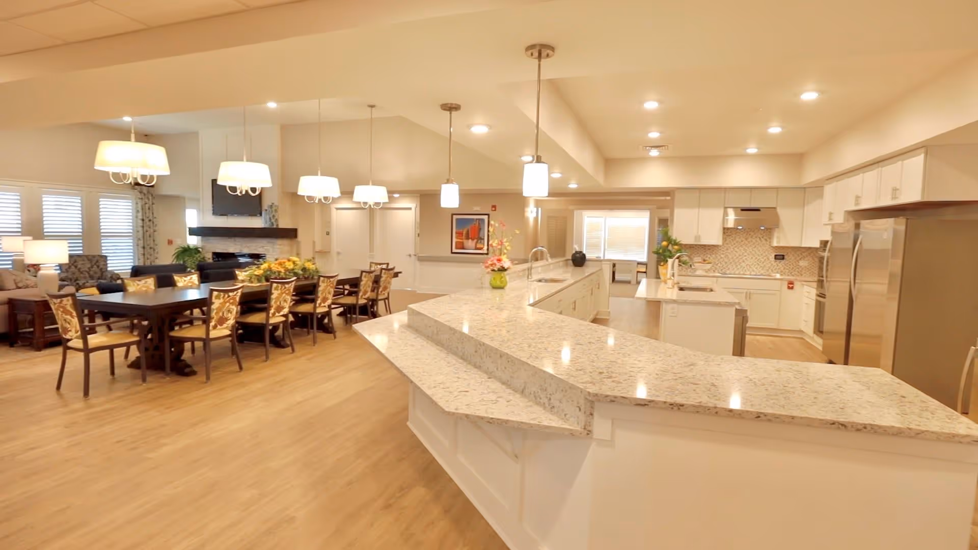 Spacious communal kitchen and dining area with a large granite island, dining table, pendant lights, and seating in an assisted living facility.