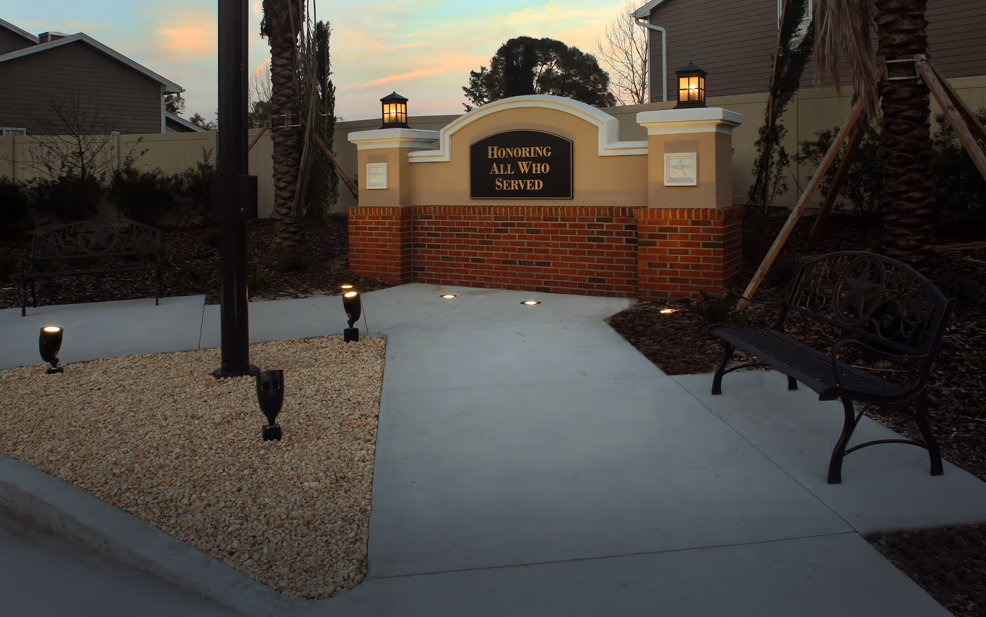 Outdoor memorial area at Anthem Lakes with a brick and stucco wall featuring a plaque that reads 'Honoring All Who Served', surrounded by palm trees, benches, and pathway lighting at dusk.