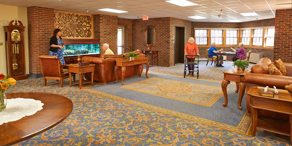 A spacious senior living facility common area with patterned carpet and brick walls. Several elderly people are present: one woman using a walker, two people sitting at a table near windows, and two others near a large fish tank. The room is furnished with leather sofas, wooden tables, a grandfather clock, and decorative wall art.