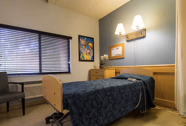 A small, neatly arranged bedroom in a senior living facility with a single bed covered in a dark blue patterned blanket. The bed has a remote control attached to it. There is a wooden nightstand with a potted plant and a framed calendar on the wall above it. A colorful hot air balloon picture hangs on the adjacent wall. The room has a window with closed blinds and a chair next to it. Two wall-mounted lamps provide lighting above the bed.