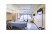 A bright and clean nursing home bedroom with a single hospital-style bed covered with a blue blanket, a large window with curtains allowing natural light, a wall-mounted television, and a small wooden chair and table near the window.