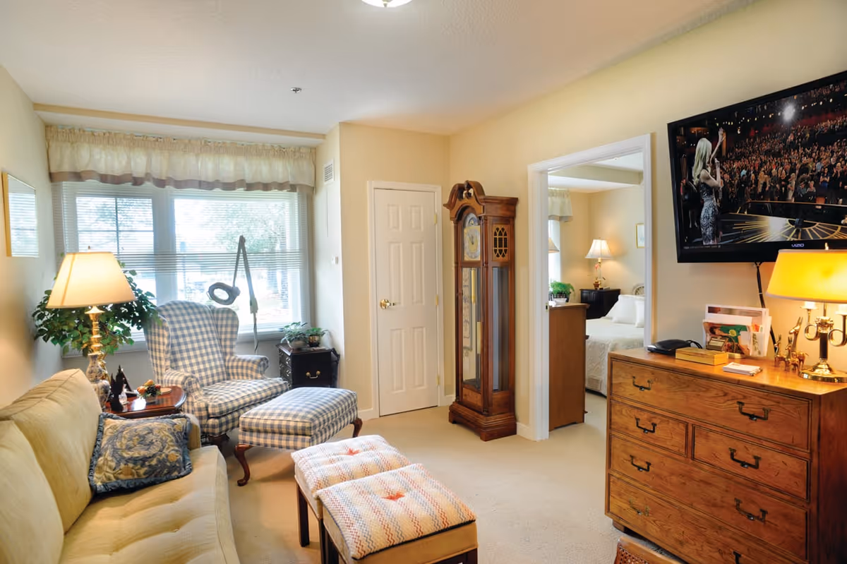 A cozy living room with a beige sofa, a blue and white checkered armchair with matching ottoman, two small cushioned stools, a wooden grandfather clock, and a wooden dresser with a lamp and a TV mounted above it. The room has a large window with blinds and a valance, and an open doorway leading to a bedroom with a bed and nightstand with a lamp.