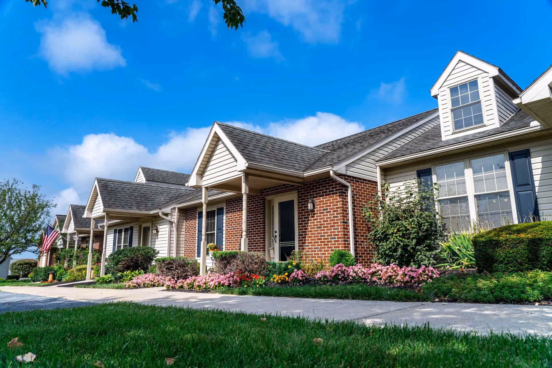 Row of single-story residential units with brick and siding exteriors, each with a small porch and well-maintained flower beds in front. The sky is clear with a few clouds, and there is a green lawn and sidewalk in the foreground.