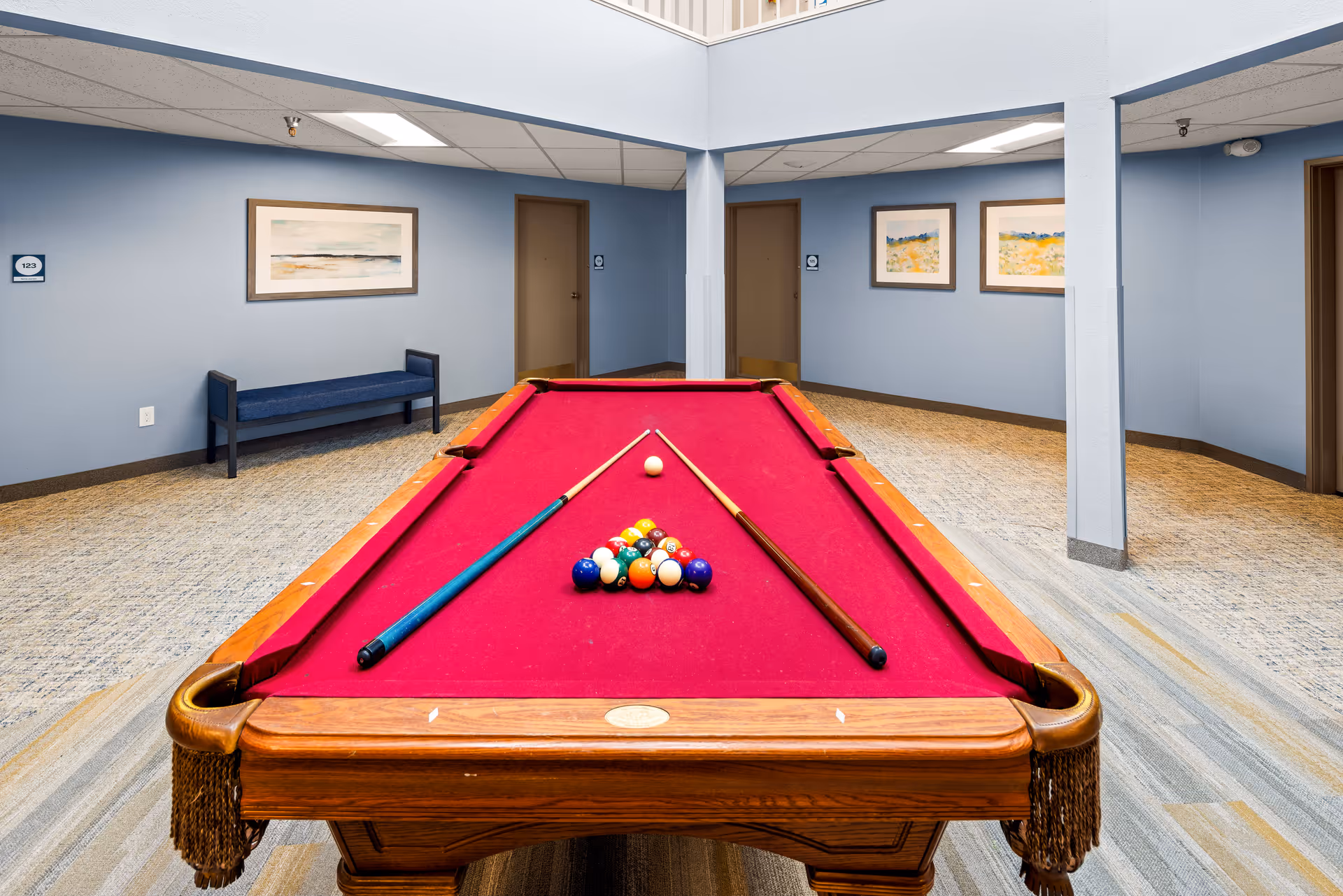 Indoor recreational room with a red felt pool table set up for a game, two pool cues resting on the table, light blue walls with framed artwork, a blue bench, and several closed doors.