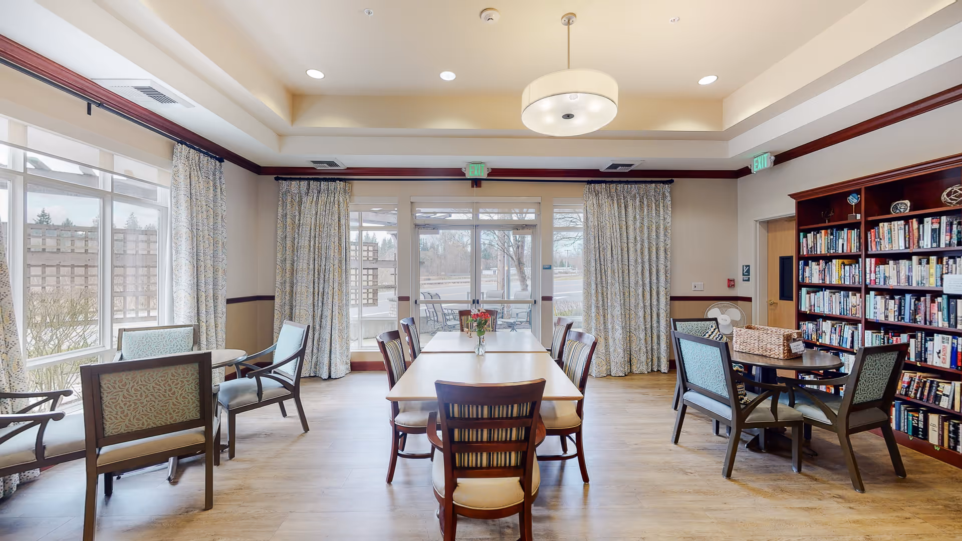 A bright and spacious common room with large windows and glass doors letting in natural light. The room features a long wooden table with chairs in the center, a round table with chairs to the right near a large bookshelf filled with books, and another small table with chairs near the windows on the left. The ceiling has recessed lighting and a modern hanging light fixture. Curtains frame the windows and doors, and the floor is light wood.