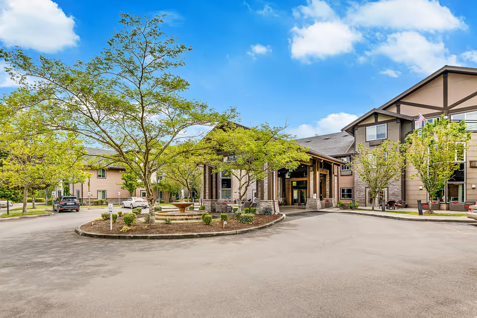 Exterior view of Meridian at Stone Creek senior living facility showing the main entrance with a covered driveway, landscaped roundabout with trees and a fountain, and a multi-story building under a blue sky with some clouds.