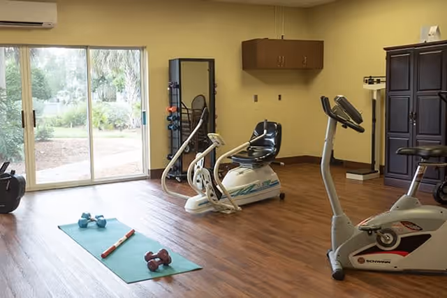 A small fitness room with exercise equipment including a stationary bike, a recumbent bike, a set of dumbbells on a green exercise mat, and a rack with various weights. The room has wooden flooring, beige walls, a large sliding glass door showing a garden outside, and a wall-mounted air conditioner.