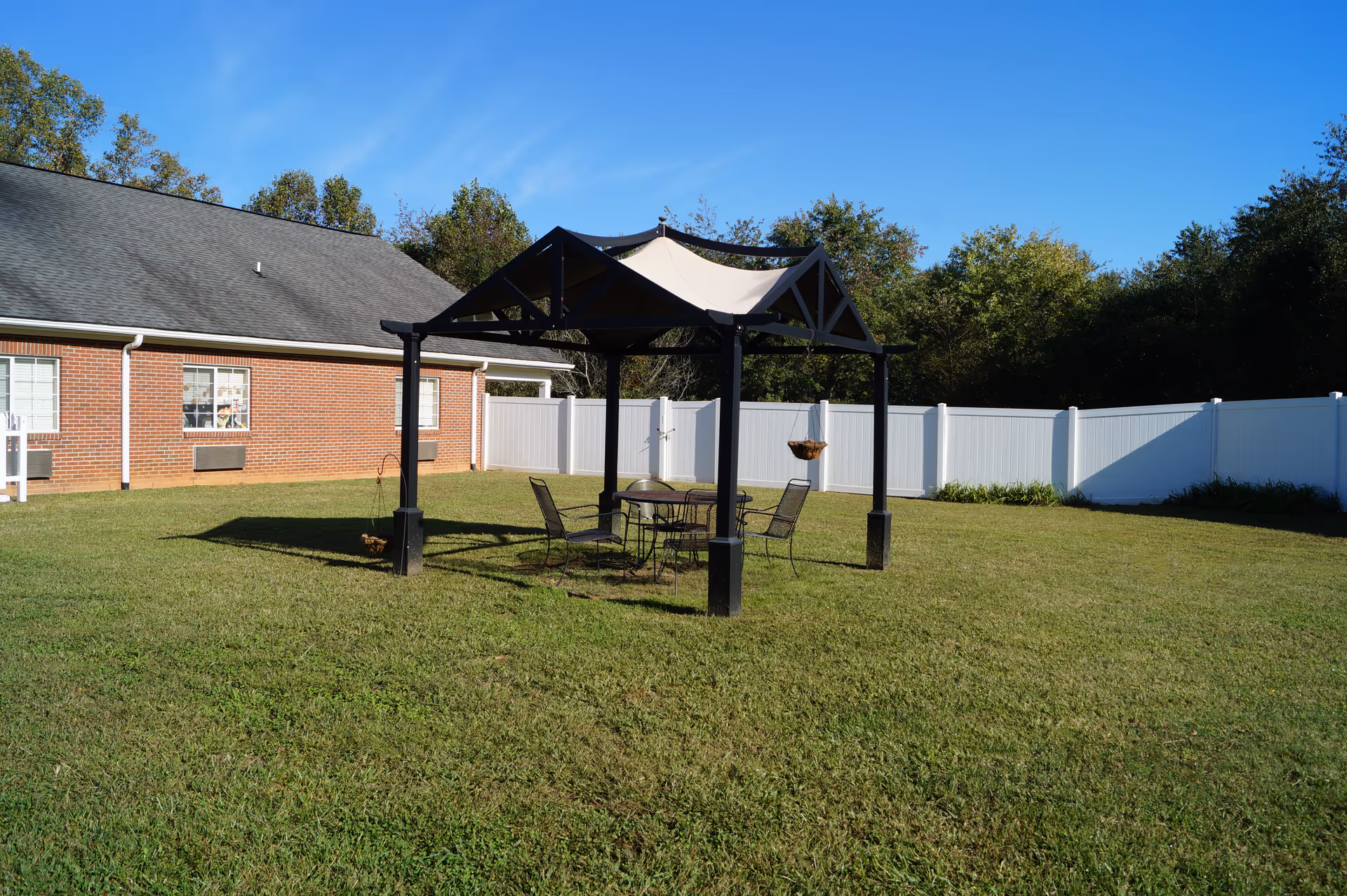 Outdoor courtyard with a black gazebo sheltering a table and chairs on a grassy lawn beside a brick building and white fence.