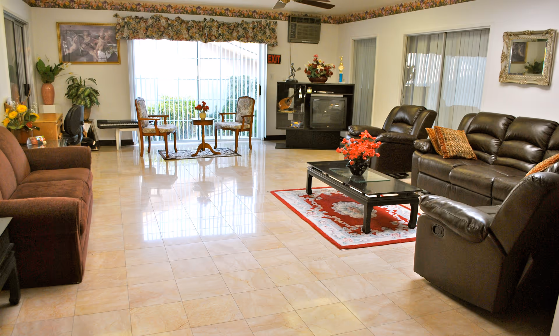 A spacious living room with a mix of brown fabric and leather sofas arranged around a black coffee table with a red floral centerpiece on a decorative rug. The room has large windows with vertical blinds and floral valances, a TV on a stand, a small table with two chairs near the window, and various plants and decorative items.
