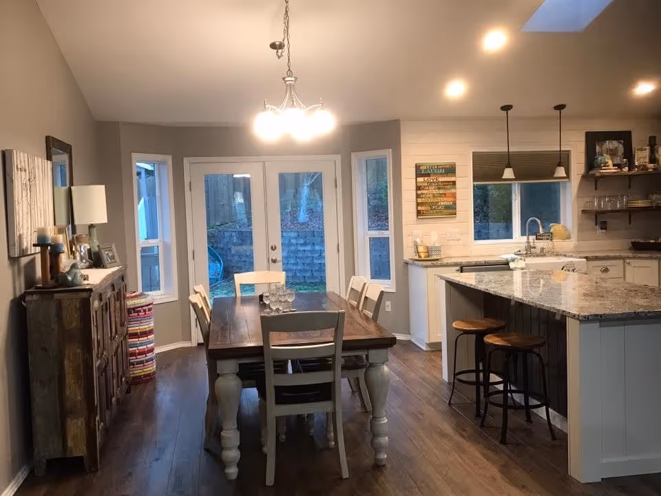 A cozy dining area and kitchen with wooden flooring. The dining area features a wooden table with six chairs and a chandelier overhead. The kitchen has a large island with a granite countertop, two bar stools, a sink, and pendant lights. There are windows and glass doors letting in natural light, and a colorful wall decoration with inspirational words is visible near the kitchen window.