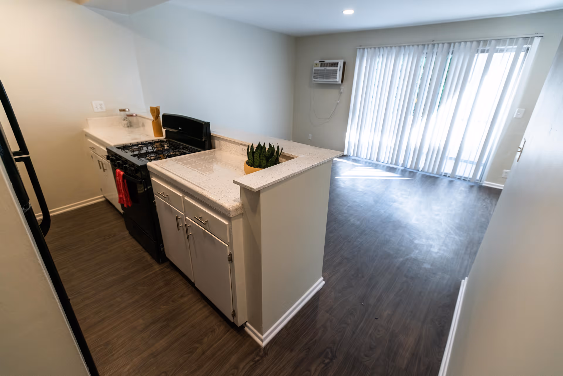 Interior view of a small kitchen area with a gas stove, white cabinets, and a countertop with a small potted plant. The kitchen opens into a living space with wood flooring and vertical blinds covering a sliding glass door. An air conditioning unit is mounted on the wall.