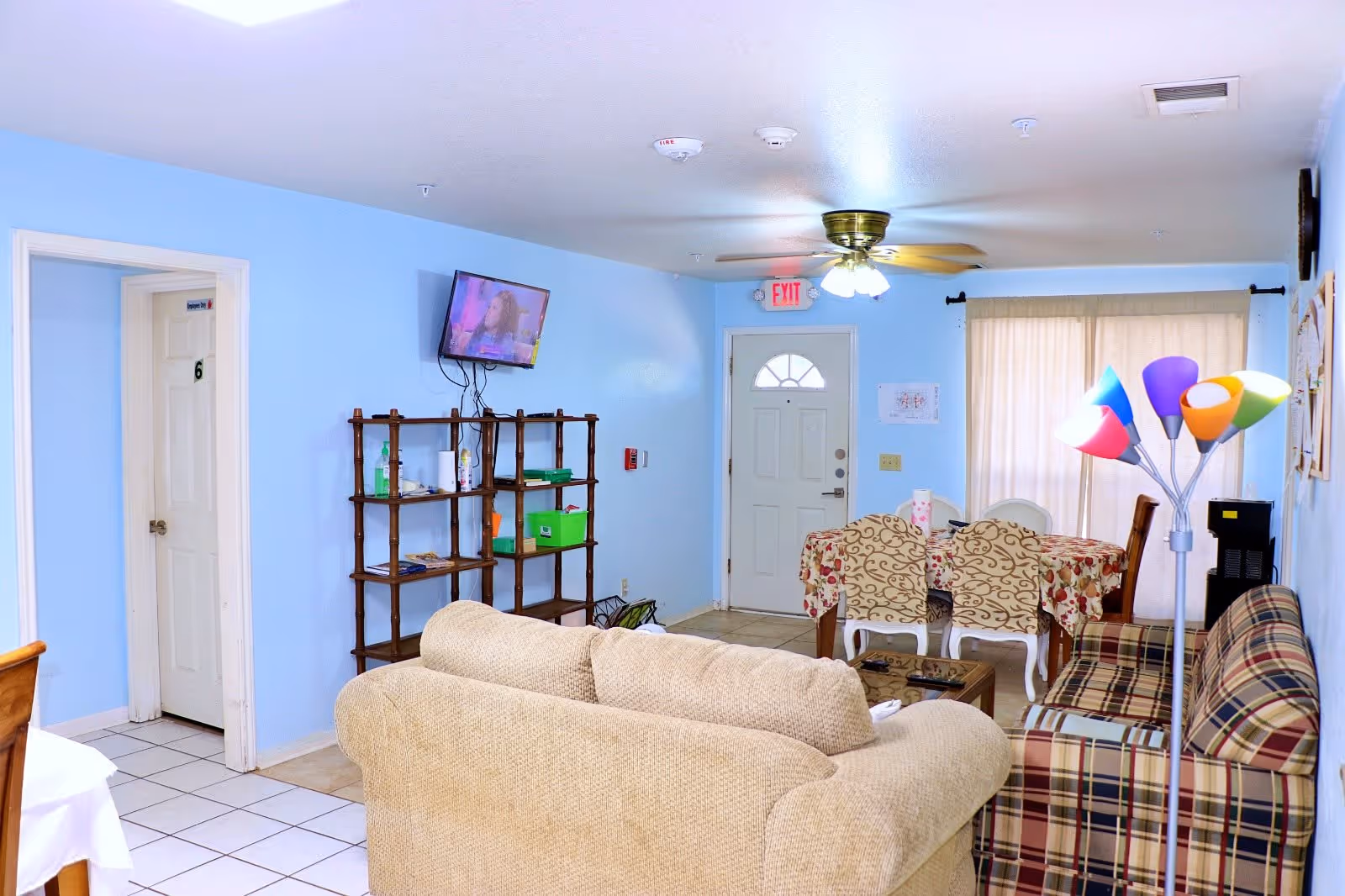 Interior view of a living room and dining area with light blue walls. The room features a beige couch, a plaid couch, a wooden shelf with various items, a wall-mounted TV, a ceiling fan with lights, a dining table with floral tablecloth and four chairs, a colorful floor lamp, and a white door with an exit sign above it. The floor is tiled in white.