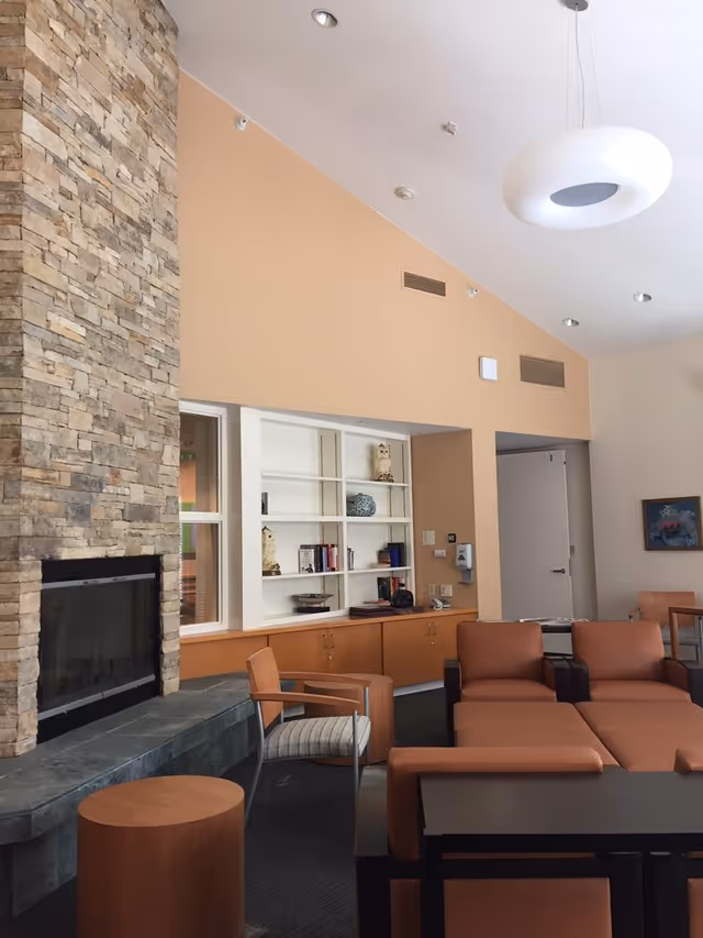 A cozy living room area in Westland House featuring a stone fireplace on the left, a built-in white bookshelf with decorative items and books, tan walls, modern ceiling light fixture, and comfortable brown leather seating with wooden side tables.