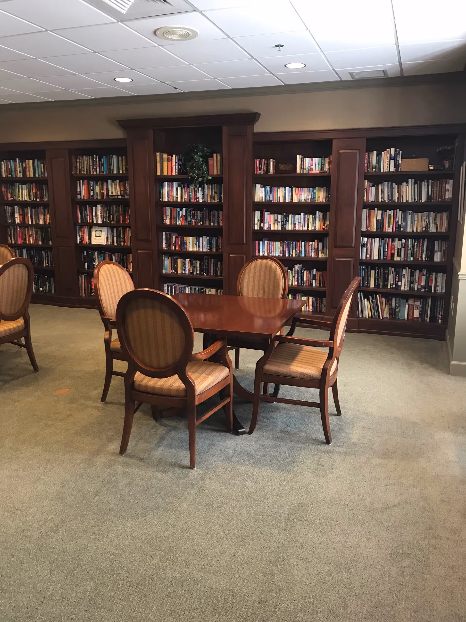 A quiet library room with wooden bookshelves filled with books along the wall. In front of the shelves, there is a wooden table surrounded by four upholstered chairs with striped fabric. The room has a carpeted floor and a white ceiling with recessed lighting.
