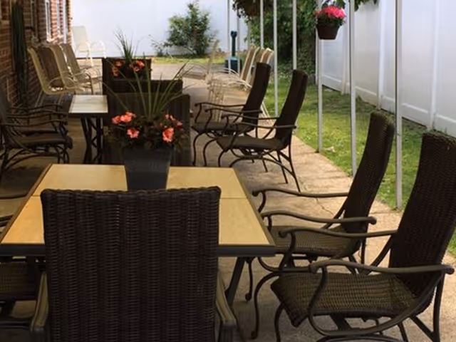 Covered outdoor patio with multiple wicker dining tables and chairs, potted plants, and a white fence.