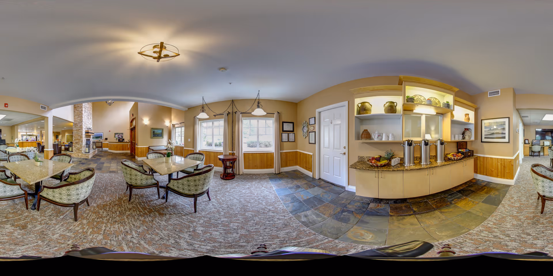 A spacious and well-lit common area in a senior living facility featuring multiple seating arrangements with tables and cushioned chairs. The room has carpeted flooring with a tiled section near a counter area that holds coffee dispensers and fruit trays. Large windows allow natural light to brighten the space, and decorative items are displayed on shelves above the counter. The area extends into another room with additional seating and a stone fireplace.